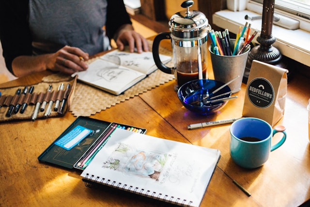 A person at a desk with pens, pencils, and a notebook, focused on a creative project.
